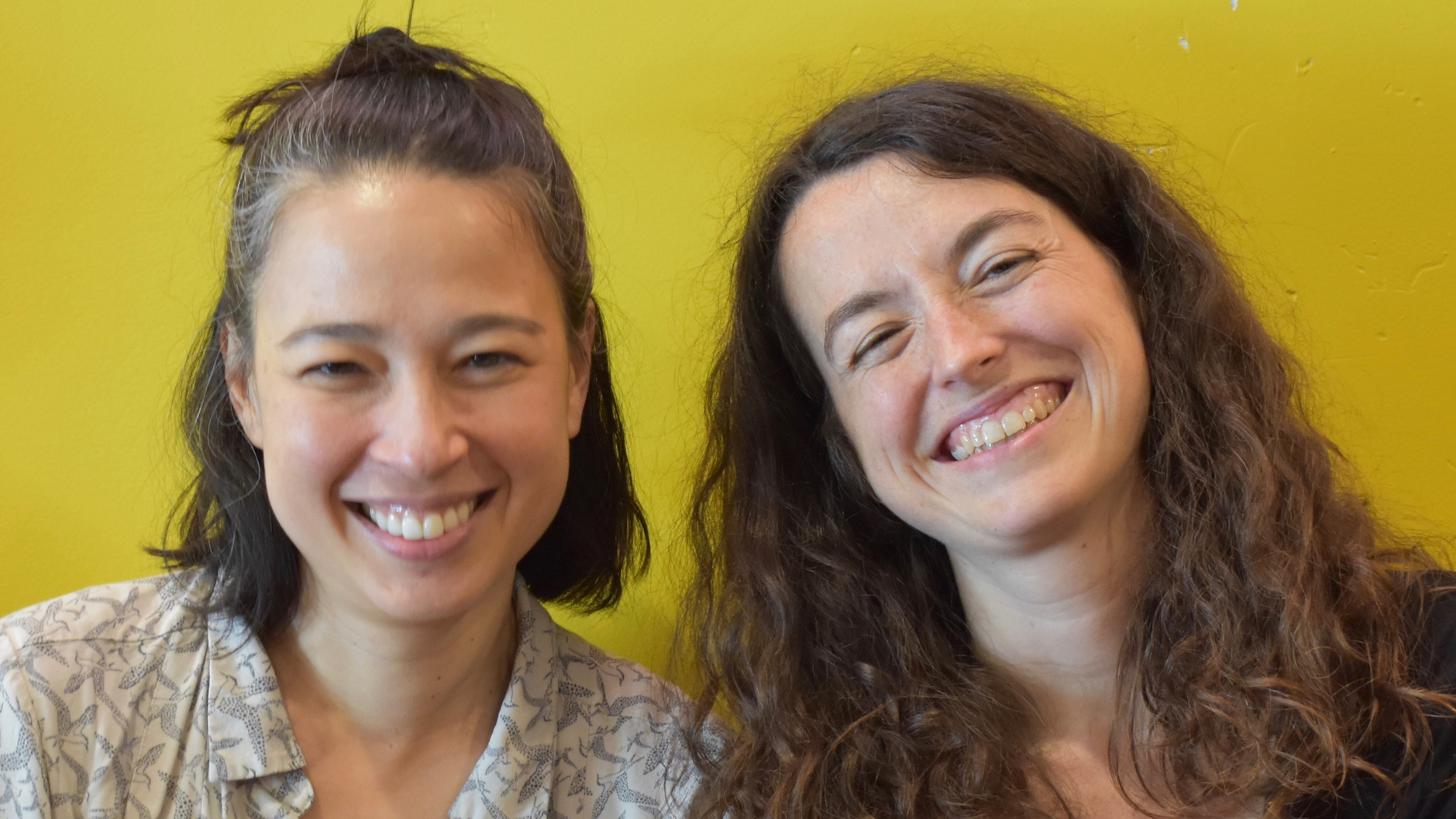 Professors Valentine Hacquard and Ellen Lau, with broadly smiling faces, sitting together in front of a bright yellow wall